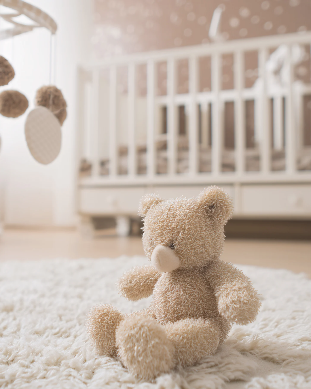 Stuffed teddy bear sitting on a nursery rug with a crib in the background.
