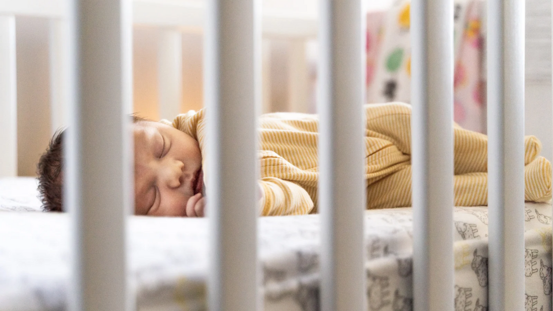 Sleeping newborn baby in a yellow outfit lying peacefully in a crib, viewed through white crib rails.