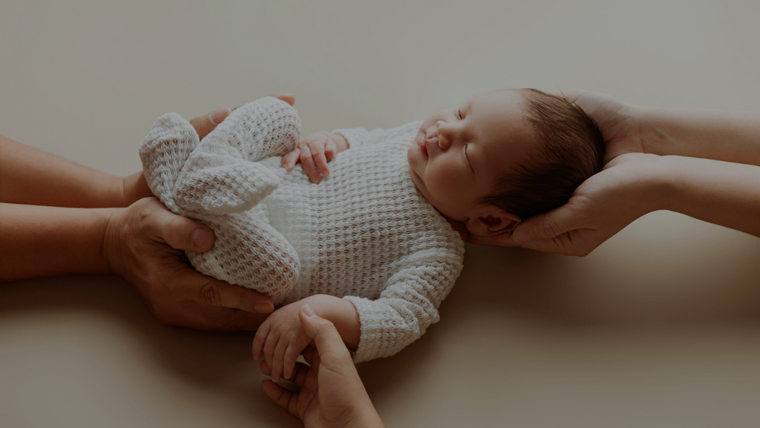 A newborn dressed in a white sleeper with hand supporting their head and body with on hand hold the baby's hand. 