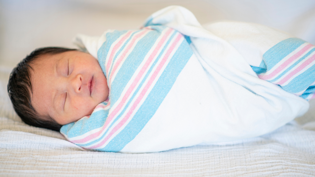 Newborn swaddled in a hospital style blanket on lying on a white sheet.