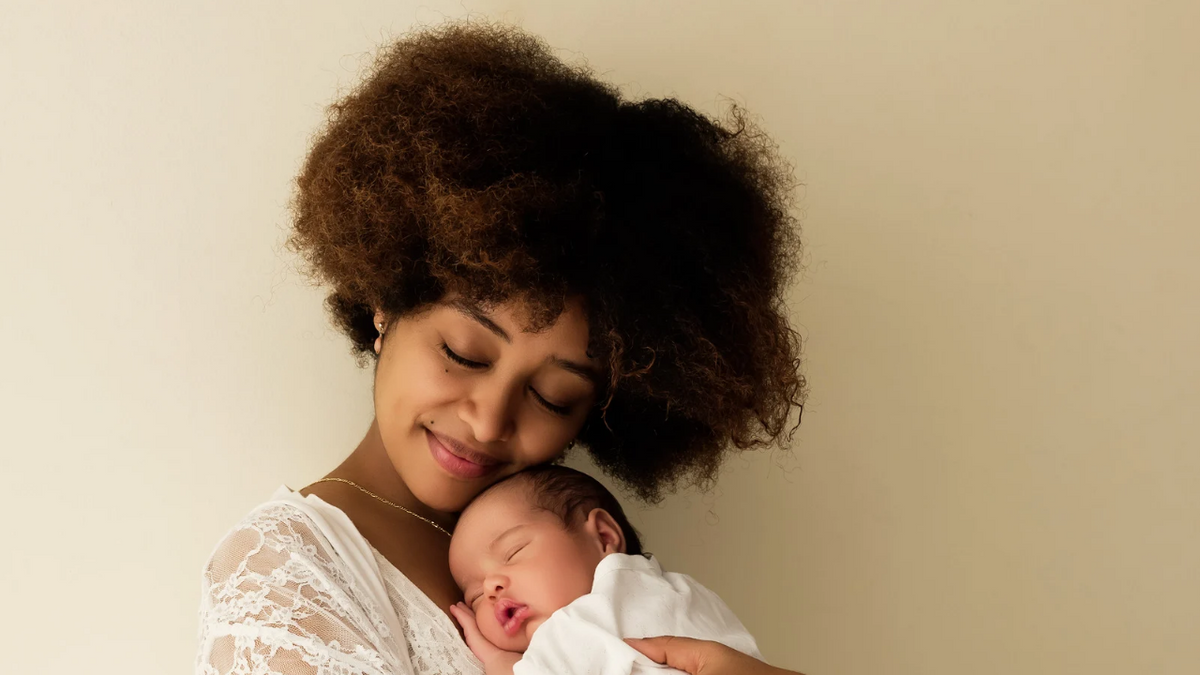 A woman with an afro hairstyle holds a sleeping newborn baby wrapped in a white swaddle against a plain beige background.