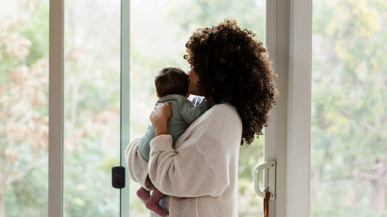 A woman with curly dark hair holds a baby in front of a large window with a soft-focus outdoor background.