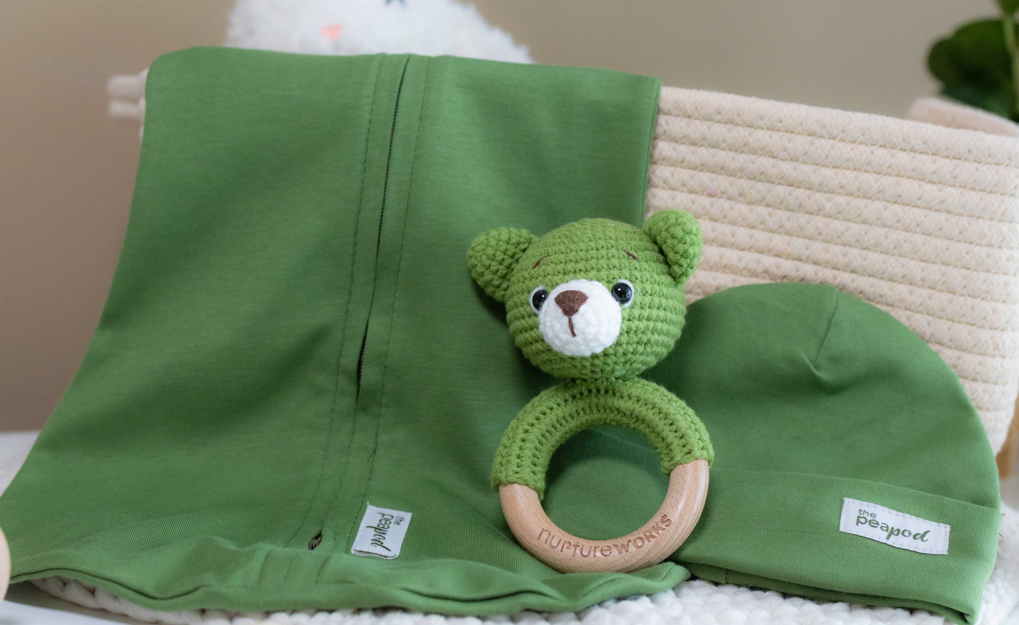 Green swaddle sleep sack with a crocheted bear rattle and hat on a neutral background