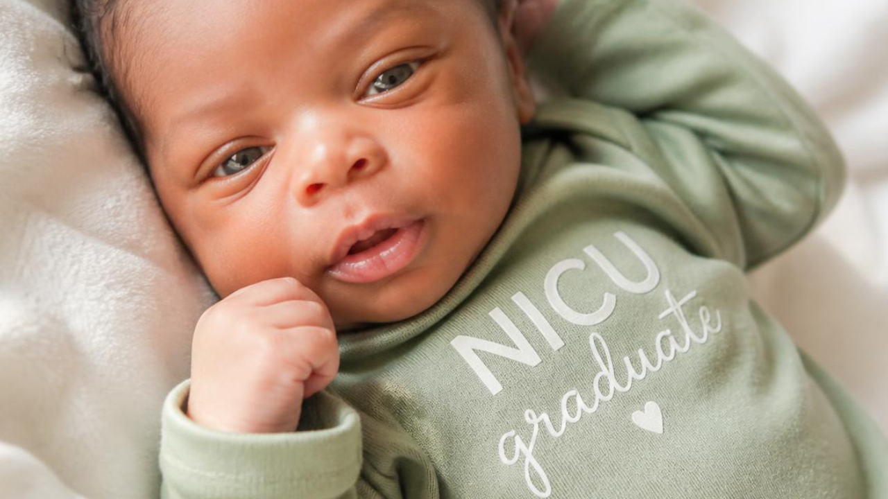 A close-up of a newborn baby wearing a sage green long-sleeve onesie with 'NICU graduate' printed in white text, lying on a soft white blanket.