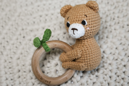 Crocheted teddy bear rattle with wooden ring on a textured surface