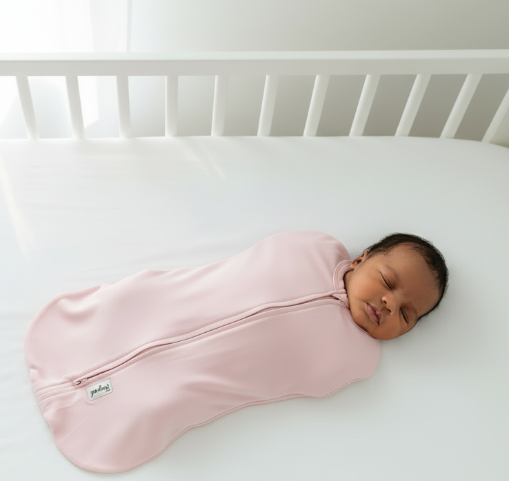 Newborn baby sleeping in a pink swaddle sleep sack in a white crib.