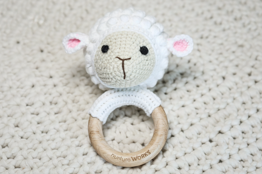 White crochet lamb rattle attached to a wooden ring on a textured beige blanket.
