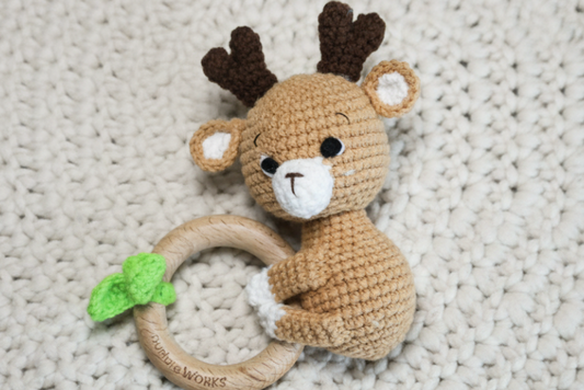 Crochet rattle with a deer design and a leaf detail, on a wooden ring on a textured blanket.  